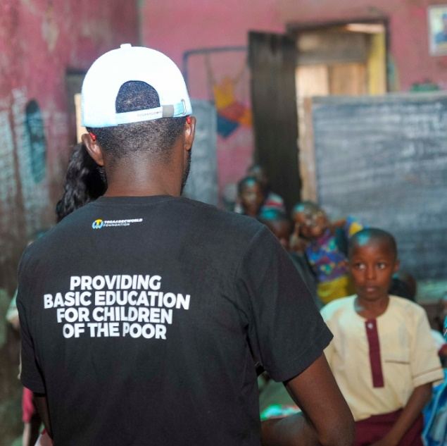 A TWF field volunteer wearing a shirt printed 'Providing basic education for children of the poor', facing a classroom of children.