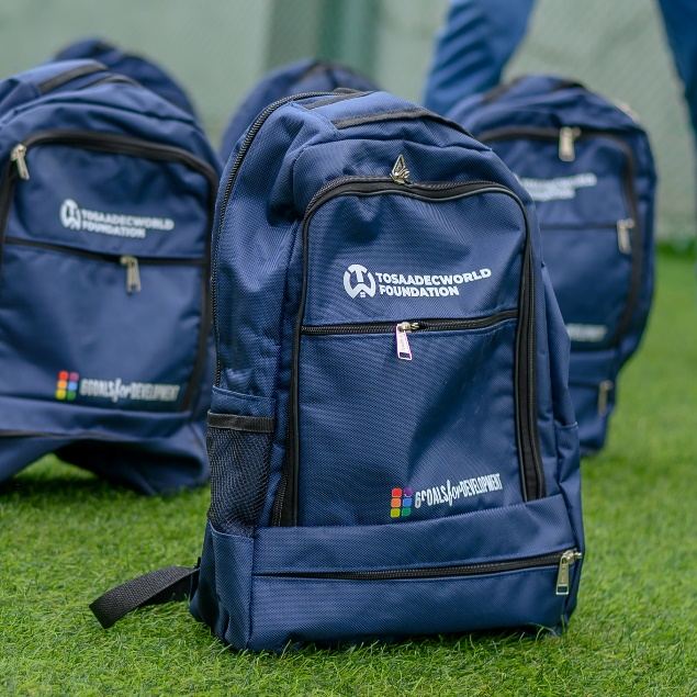 Navy blue TosaadecWorld Foundation backpacks lined up on grass, ready for distribution to sponsored children.